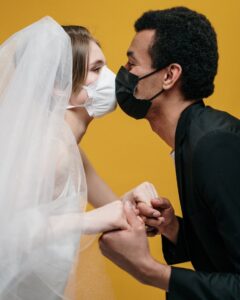 A bride and groom both wearing surgical masks holdings hands and kissing