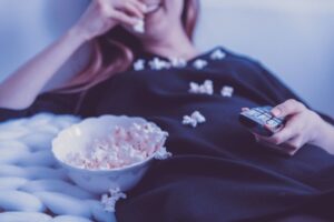 A girl relaxing eating popcorn and holding a remote flipping through channels on television, a pleasant activity to treat oneself